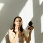 About 1 Asian woman applying makeup while sitting indoors, illuminated by natural light and shadows.